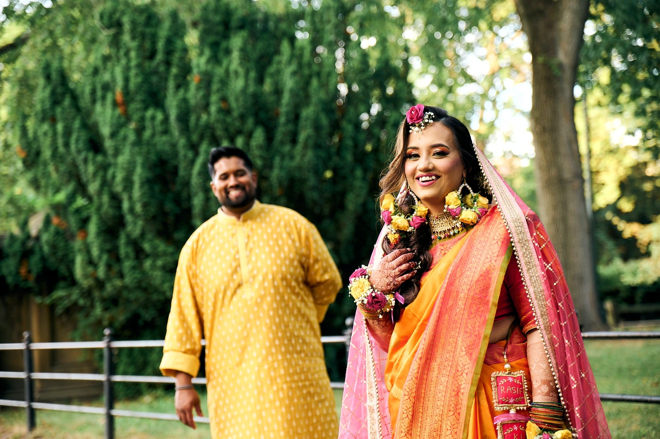 Bride in orange and pink sari with floral jewelry stepping forward, groom in yellow kurta behind smiling